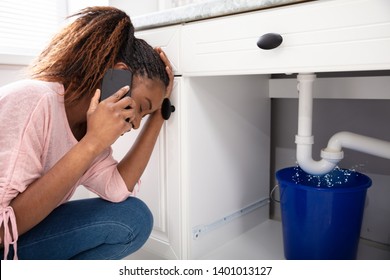 Young Woman Calling Plumber Crouching In Front Of Water Leaking From Sink Pipe