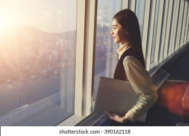 Young woman candidate is holding laptop computer, while standing in office interior and looking out of big window with city view. Female manager with net-book in hand ponder about new business project