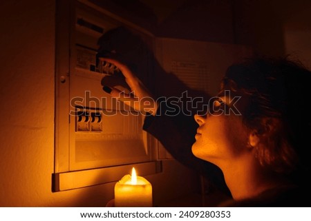 young woman with candles in complete darkness examines fuse box or electrical distribution board at home during power outage. Blackout, no electricity