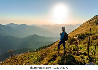 young woman in cap with backpack standing among Caucasus mountains and looking at setting sun and mountain view. Landscape, Aibga ridge, hiking, travel alone, beauty in nature,  World Tourism Day