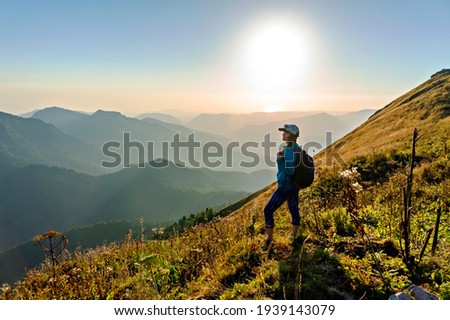 young woman in cap with backpack standing among Caucasus mountains and looking at setting sun and mountain view. Landscape, Aibga ridge, hiking, travel alone, beauty in nature,  World Tourism Day