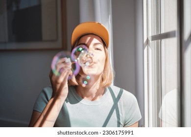 Young woman in cap blowing soap bubbles next to the window. Concept: fun, house, objects