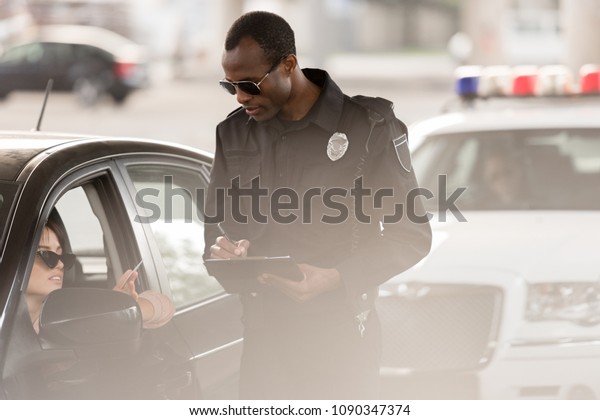 young woman in car giving driver\
license to african american policeman with clipboard and\
pen