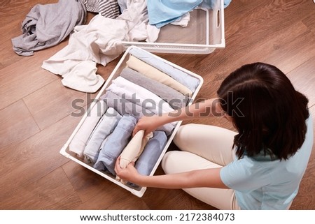A young woman carefully puts stacks of clothes in a metal laundry basket. The concept of restoring order, cleaning, cluttering, organizing space. 