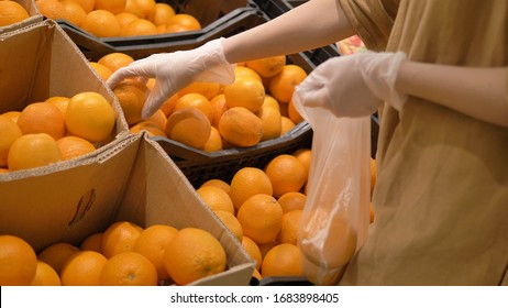 A Young Woman Carefully Selects Fresh Oranges In A Supermarket In Rubber Protective Gloves And Puts Them In A Plastic Bag. Personal Protective Equipment Against Coronavirus.