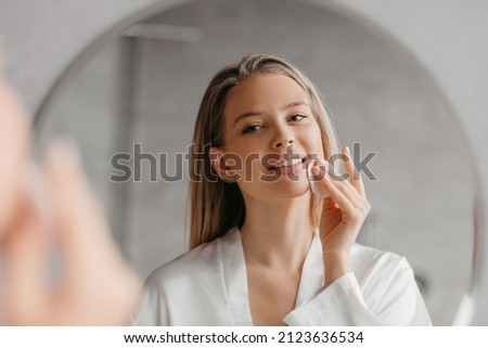 Young woman caring for facial skin using cotton pad and looking in round mirror in bathroom interior. Beauty care and pampering. Daily female skincare routine concept