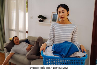 Young woman carrying a basket of dirty clothes doing chores at home while lazy man relaxing and sleeping on sofa couch. Couple having a housework domestic problem - Shutterstock ID 1864837282