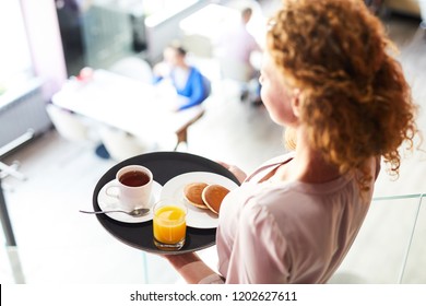 Young Woman Carrying Pancakes, Tea And Juice On Tray While Moving To One Of Tables In Cafe