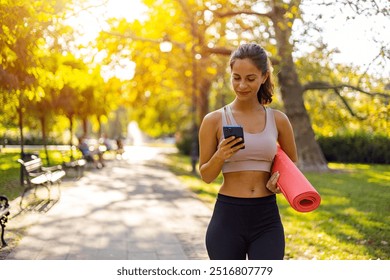 A young woman carrying a yoga mat and using a smartphone while walking in a sunny park. Embracing healthy outdoor activities and digital connectivity. - Powered by Shutterstock