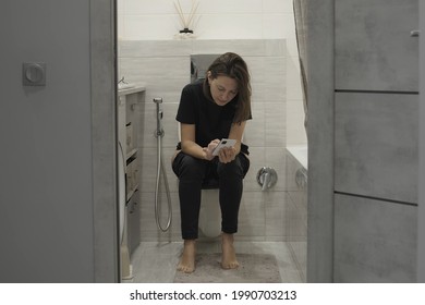 Young woman in casual clothes in bathroom, sitting on toilet bowl and using cellphone. Woman On The Toilet Looking At Her Mobile Phone - Powered by Shutterstock