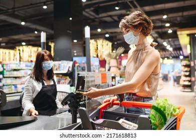 Young Woman In Casual Clothes Mask Shopping At Supermaket With Grocery Cart Put Mobile Phone To Modern Bank Payment Terminal To Process Acquire Credit Payments At Checkout Near Cashier Inside Store.