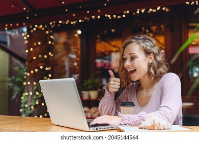 Young Woman In Casual Clothes Sit Alone At Table In Cafe Shop Eat Breakfast Hold Use Work On Laptop Pc Computer Talk By Viceo Call Show Thumb Up Drink Coffee Relax In Restaurant In Free Time Indoors.