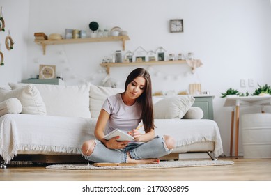 Young Woman In Casual Clothes Sitting On The Floor. The girl makes notes in a notebook. Cozy Home Interior. Scandinavian Kitchen Interior.