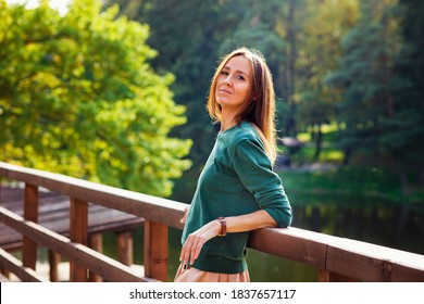 A young woman in casual clothes stands on a bridge in a Park and looks at the camera. Happy cheerful woman enjoying the weekend and the good weather outdoors.