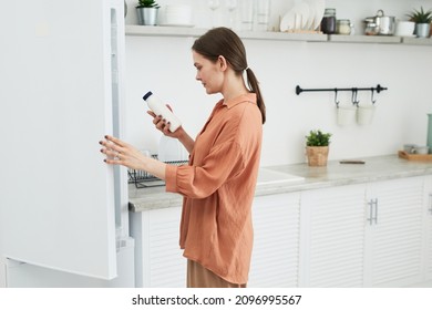 Young Woman In Casual Clothing Reading Label On The Bottle Opening The Fridge During Her Preparation In The Kitchen