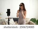 A young woman in casual home attire energetically dances and sings in front of a phone and ring light in a bright, stylish living room. She strikes various expressive poses while performing, capt