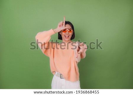 Young woman in casual peach sweater and orange glasses isolated on green olive background pointing at you mockery fool day showing horn symbol copy space