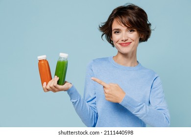 Young woman in casual sweater point finger on pressed juice green orange vegetable smoothie as detox diet isolated on plain pastel light blue background studio portrait. People lifestyle food concept