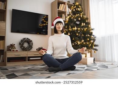 Young woman of Caucasian descent sits in lotus position meditating wearing Santa hat near Christmas tree. Scene captures peaceful holiday ambiance, blending relaxation and festive decor. - Powered by Shutterstock