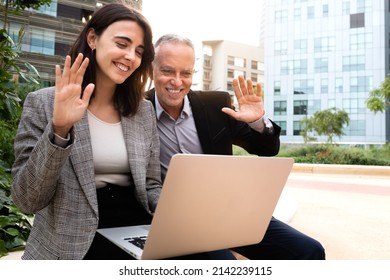 Young Woman And Caucasian Man Wave Hello On Business Video Call Sitting On A Bench Outside Office Buildings. Copy Space.