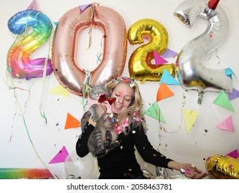 young woman celebrating new year 2022 at home quarantine. smiling girl holding cute british cat in shirt. big number balloons. silvester party. december 31. lockdown. party decoration.confetti falling