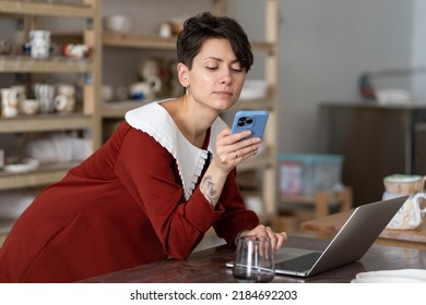 Young Woman Ceramic Artisan Using Social Media To Communicate With Customers While Standing At Desk With Laptop In Pottery Studio Or Workshop. Craft Business Owner With Mobile Phone In Ceramics Shop