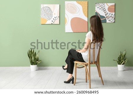 Young woman in chair near paintings on green wall at art gallery exhibition, back view