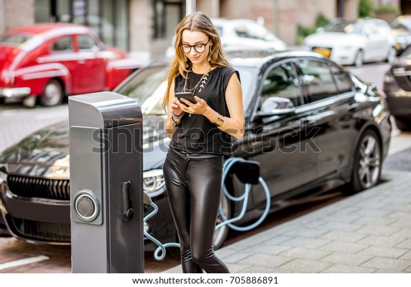 Young woman charging\
electric car standing with smart phone outdoors on the street in\
Rotterdam city