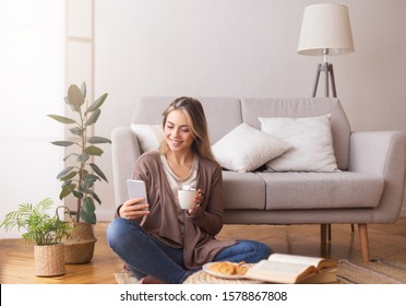 Young Woman Chatting With Friends On Cellphone, Enjoying Weekend At Home With Coffee And Book, Empty Space