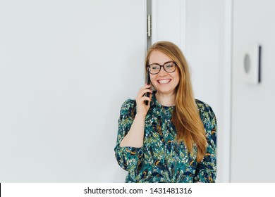 Young Woman Chatting On Her Mobile With A Smile Of Pleasure As She Leans Against A Door Indoors At Home With Copy Space On White Wall