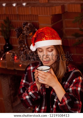 young woman in checkered shirt and red Santa hat with a cup of hot chocolate with marshmallows near the red brick fireplace. Christmas winter time