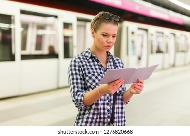 Young Woman In Checkered Shirt Waiting For Train At Subway Station, Looking At Booklet