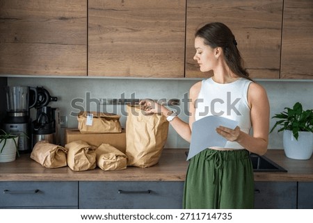 Young woman checking a list while looking at unpacked meal kit ingredients organized by recipe. Concept of planning weekly cooking using convenient pre-portioned grocery delivery