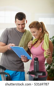 Young Woman Checking Training Plan With Her Personal Trainer Standing Beside Exercise Bike At Home.
