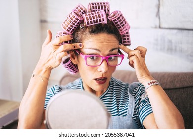 Young woman checking wrinkles near eye while applying curlers on hair and looking in front on mirror sitting on sofa in living room at home. Beautiful woman in eyeglasses getting ready with make up.