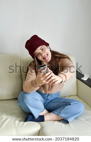 A young woman in a cheerful mood grimaces, sticks out her tongue and squints slyly, takes a selfie on the phone, has a wireless earpiece in her ear, listens to music, talks with friends.