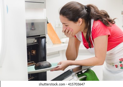 Young Woman Chef Looking Into Oven With Frustrated Facial Expression, Holding Black Burnt Bread On Tray