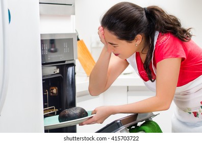 Young Woman Chef Looking Into Oven With Frustrated Facial Expression, Holding Black Burnt Bread On Tray