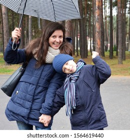 A Young Woman With A Child Under An Umbrella Laughs. Girl With A Little Boy Smiling, Walking In The Park.  The Unexpected Rain.