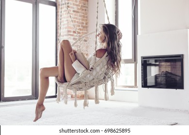 Young woman chilling at home in comfortable hanging chair near fireplace. Girl relaxing and reading book in swing in loft living room with brick walls.