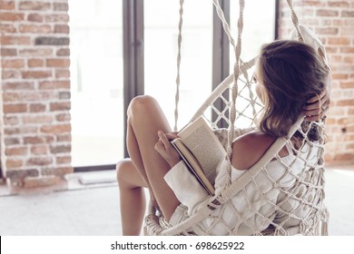 Young woman chilling at home in comfortable hanging chair. Girl relaxing and reading book in swing in loft living room with brick walls. - Powered by Shutterstock