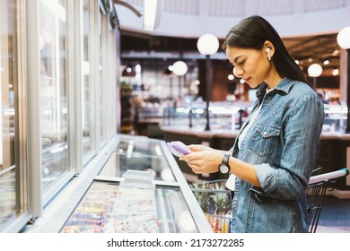 Young Woman Chooses Groceries In Supermarket Standing Near The Freezer And Refrigerators Department