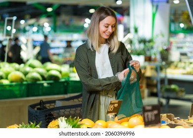 Young Woman Chooses And Picks In Eco Bag Vegetables Or Fruits Oranges In The Supermarket. Female Customer Standing A Grocery Store Near The Counter Buys And Throws In A Reusable Package In Market