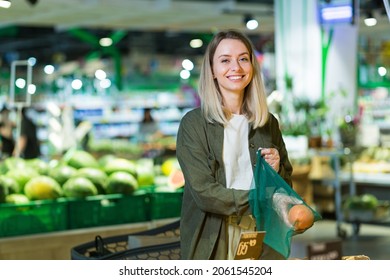 Young Woman Chooses And Picks In Eco Bag Vegetables Or Fruits Oranges In The Supermarket. Female Customer Standing A Grocery Store Near The Counter Buys And Throws In A Reusable Package In Market