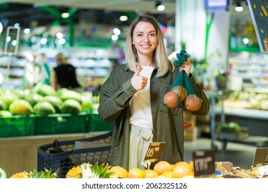 Young Woman Chooses And Picks In Eco Bag Vegetables Or Fruits Oranges In The Supermarket. Female Customer Standing A Grocery Store Near The Counter Buys And Throws In A Reusable Package In Market