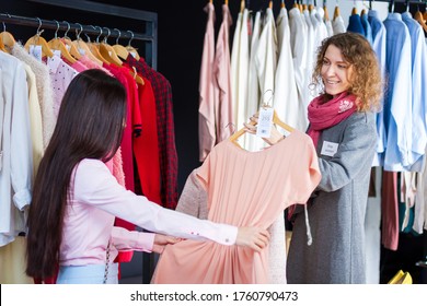 Young Woman Is Choosing, Buying Fashionable Dress In Store, Boutique In Center, Mall. Kind Seller, Consultant, Stylist, Shop Assistant Is Serving Girl. Black Friday. Shopping And Sale Concept.