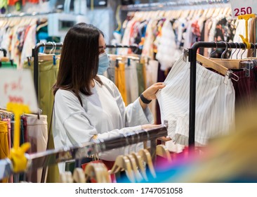 Young Woman Choosing Clothes At Shopping Mall And Her Wearing Medical Mask For Prevention From Coronavirus (Covid-19) Pandemic. New Normal Concepts