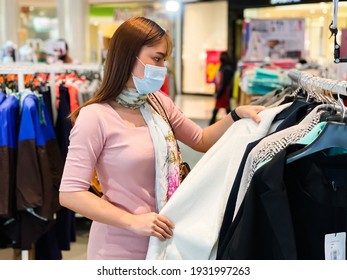 Young Woman Choosing Clothes At Shopping Mall And Her Wearing Medical Mask For Prevention From Coronavirus (Covid-19) Pandemic. New Normal Concepts