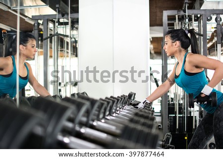 Young woman choosing dumbbells in gym. She stay near a mirror and see her reflection.