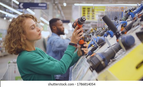 Young Woman Choosing Electric Screwdriver In Hardware Store. Side View Of Pretty Lady Examining Electric Drill In Construction Equipment Department Of Diy Shop